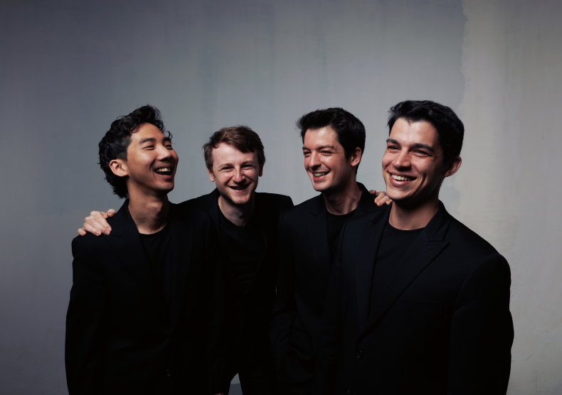 Four men in all black smile with their arms around each others' shoulders