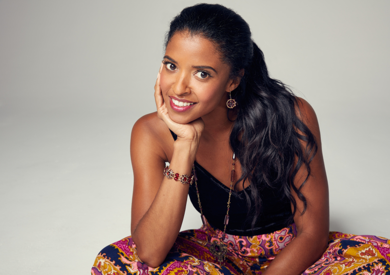 A woman sits cross-legged with her head in her hand, wearing colorful pants and a black top
