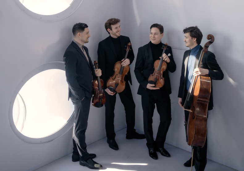 Four men of a string quartet dressed in all black hold their string instruments in a white room with circular windows