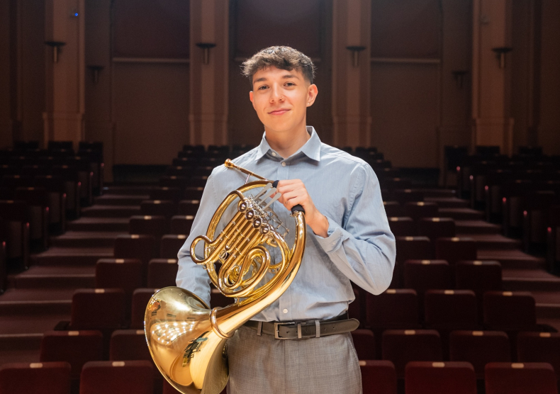 A man holding a french horn stands in an empty hall in front of the seats