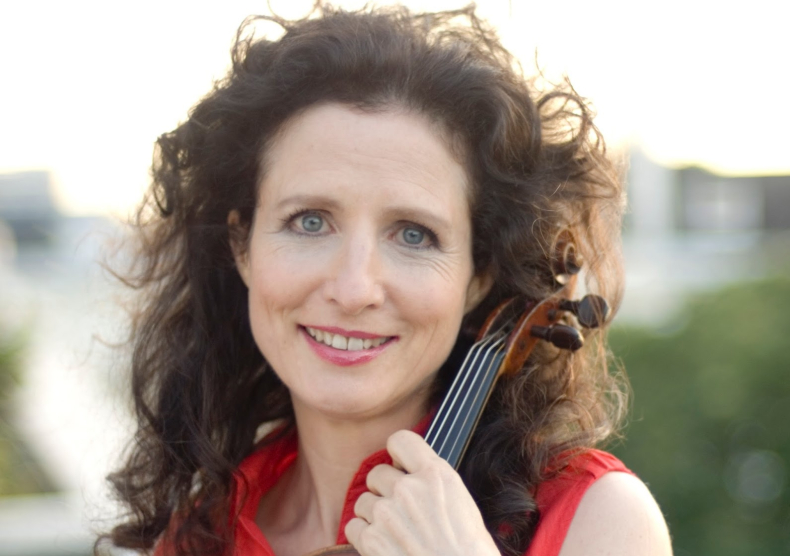 A woman with dark brown hair and wearing a red sleeveless shirt holds her violin and smiles at the camera 