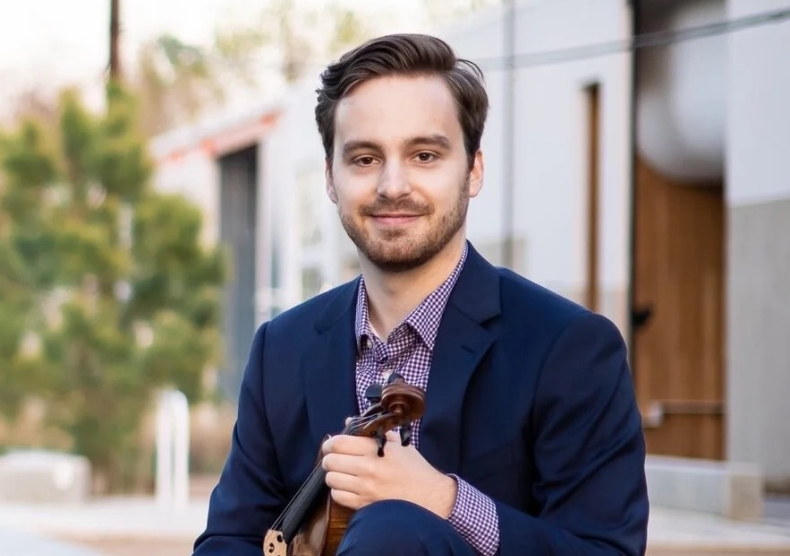 A man in a purple dress shirt and blue suit jacket sits with his violin while smiling at the camera