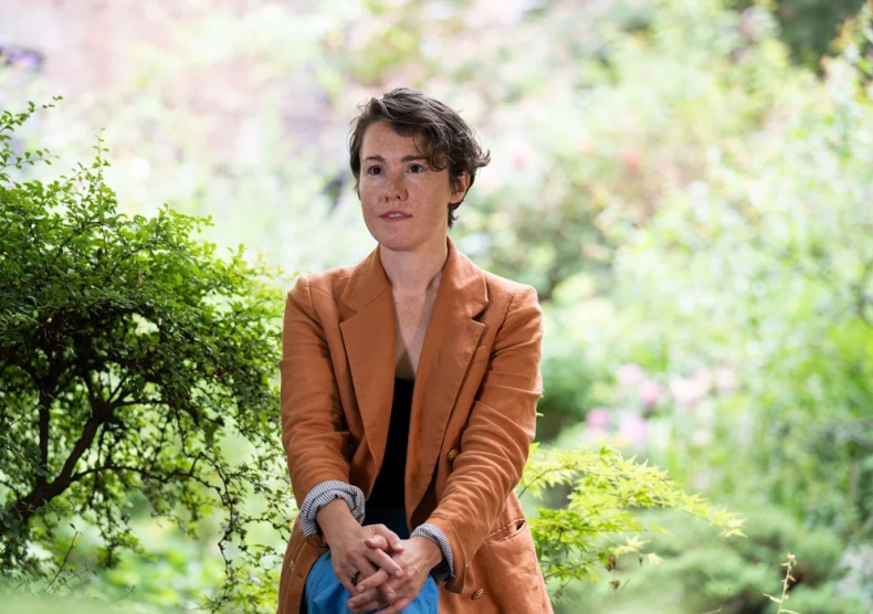 A person with an orange jacket sits with her legs crossed in front of greenery