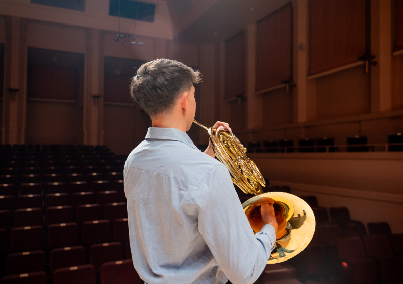 A french horn player wearing a light blue dress shirt performs in an empty recital hall