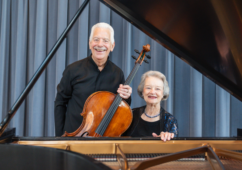 A man holding a cellist and a woman seated in front of a piano smile